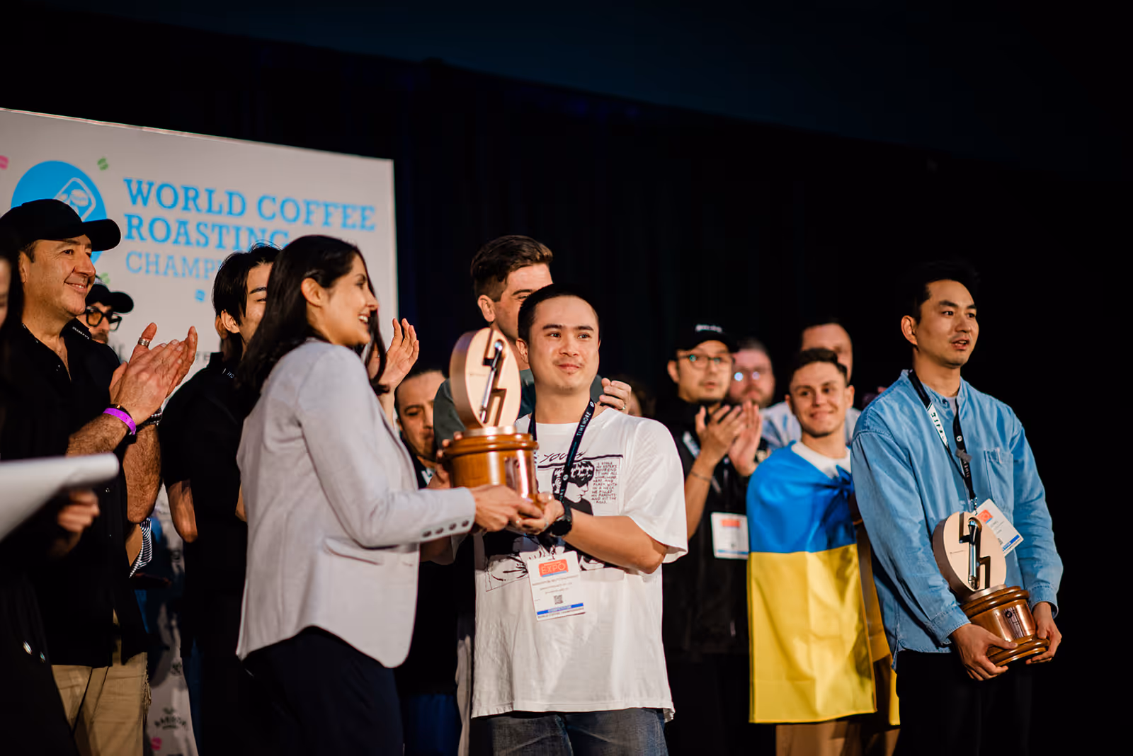 Man in white shirt receives a trophy at the World Coffee Roasting Championship while others applaud.