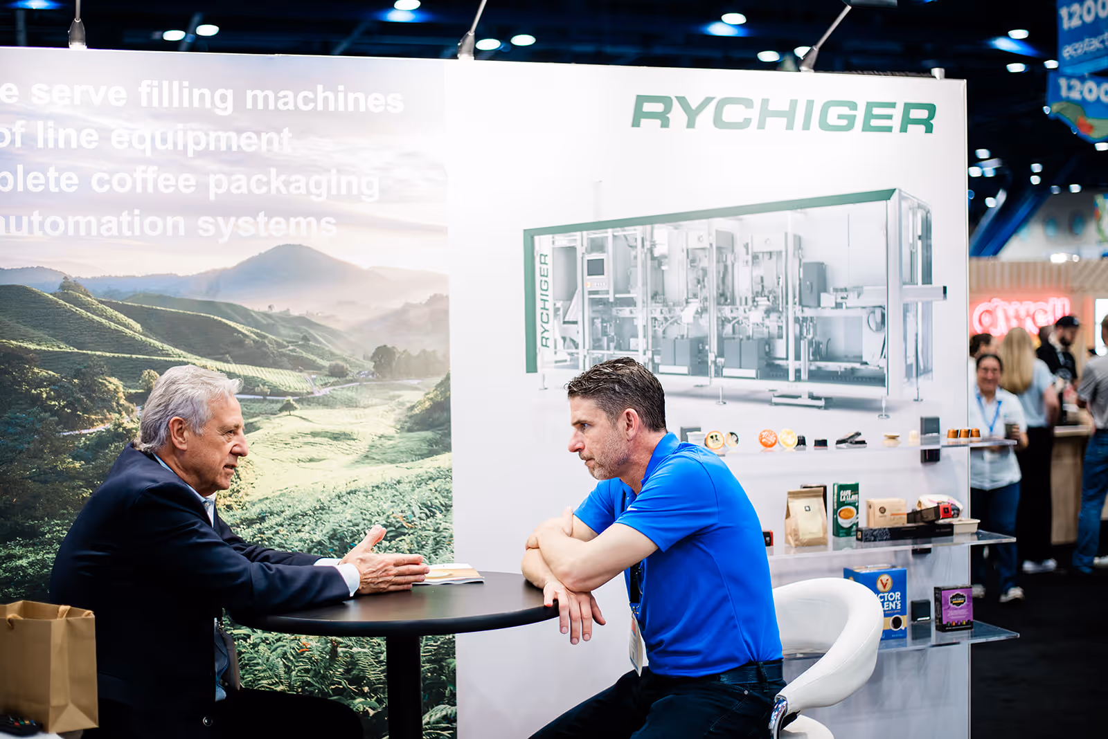 Two men seated at a round table engaged in conversation at a trade show booth with Rychiger machinery and coffee packaging displayed.
