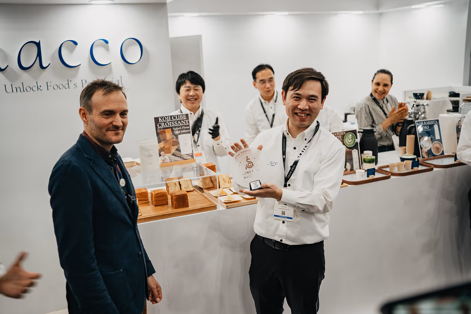 Smiling man in white shirt holding a round award plaque at a booth with baked goods and drinks, surrounded by four other people applauding or smiling.