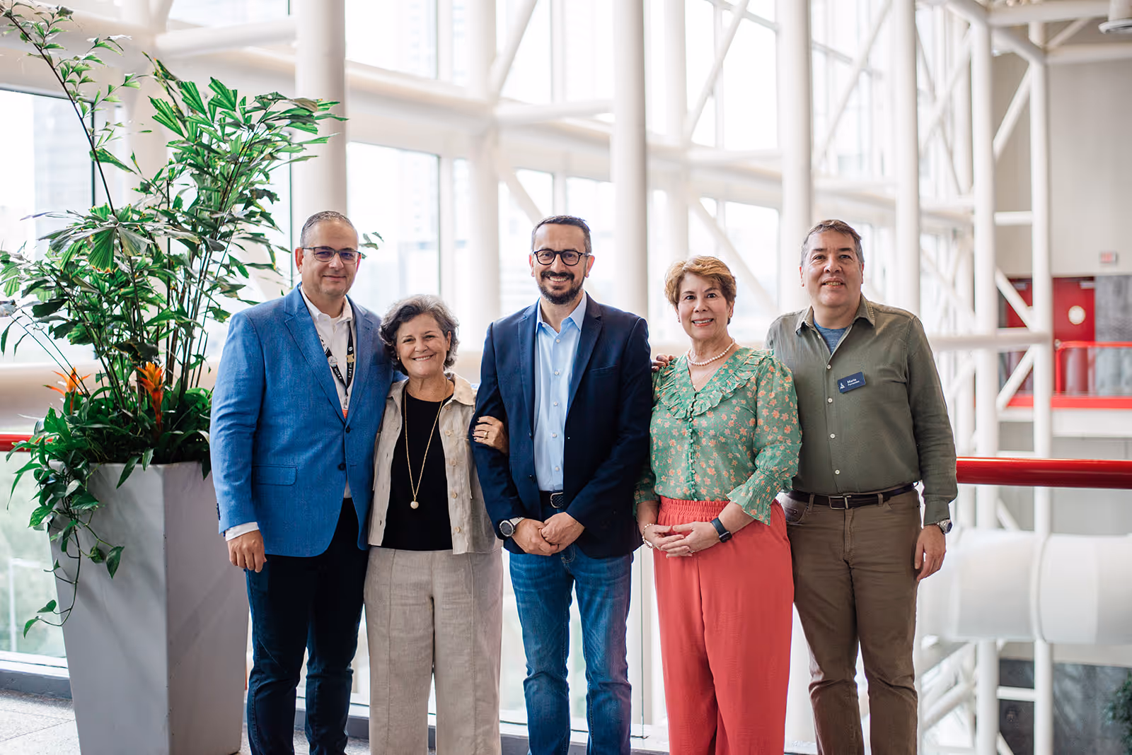 Group of five adults standing indoors by large windows, smiling at the camera, with one large potted plant on the left.