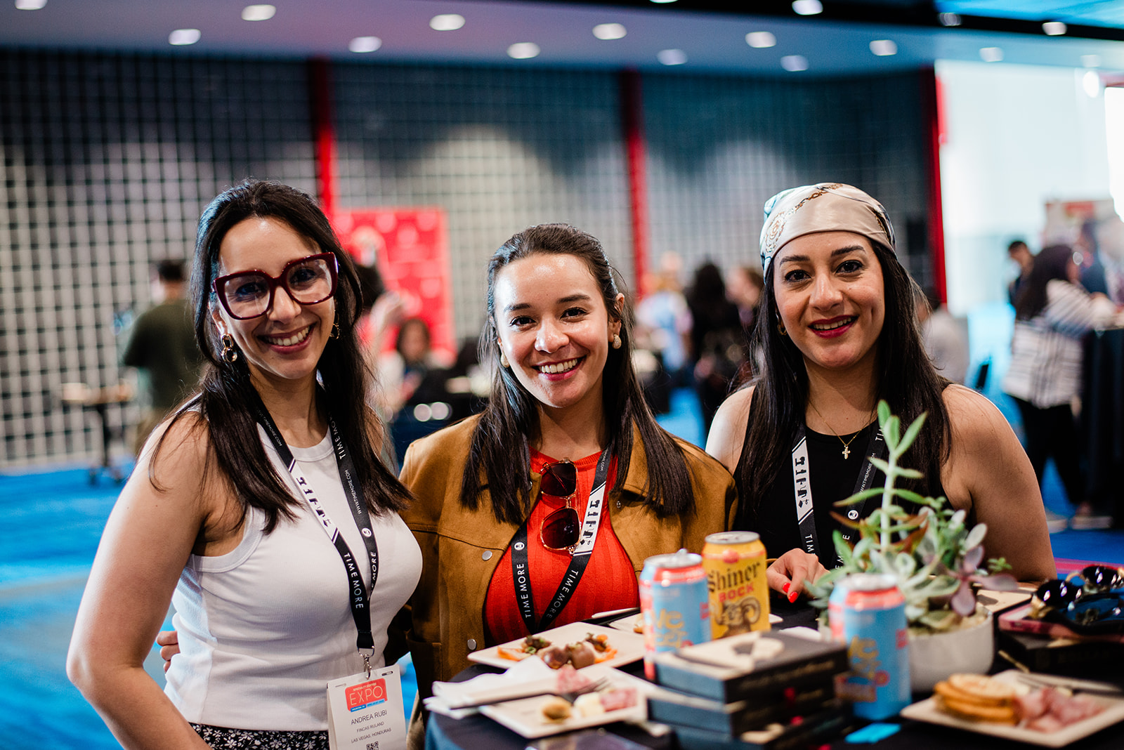 Three smiling women standing together at a social event with food and drinks on a nearby table.