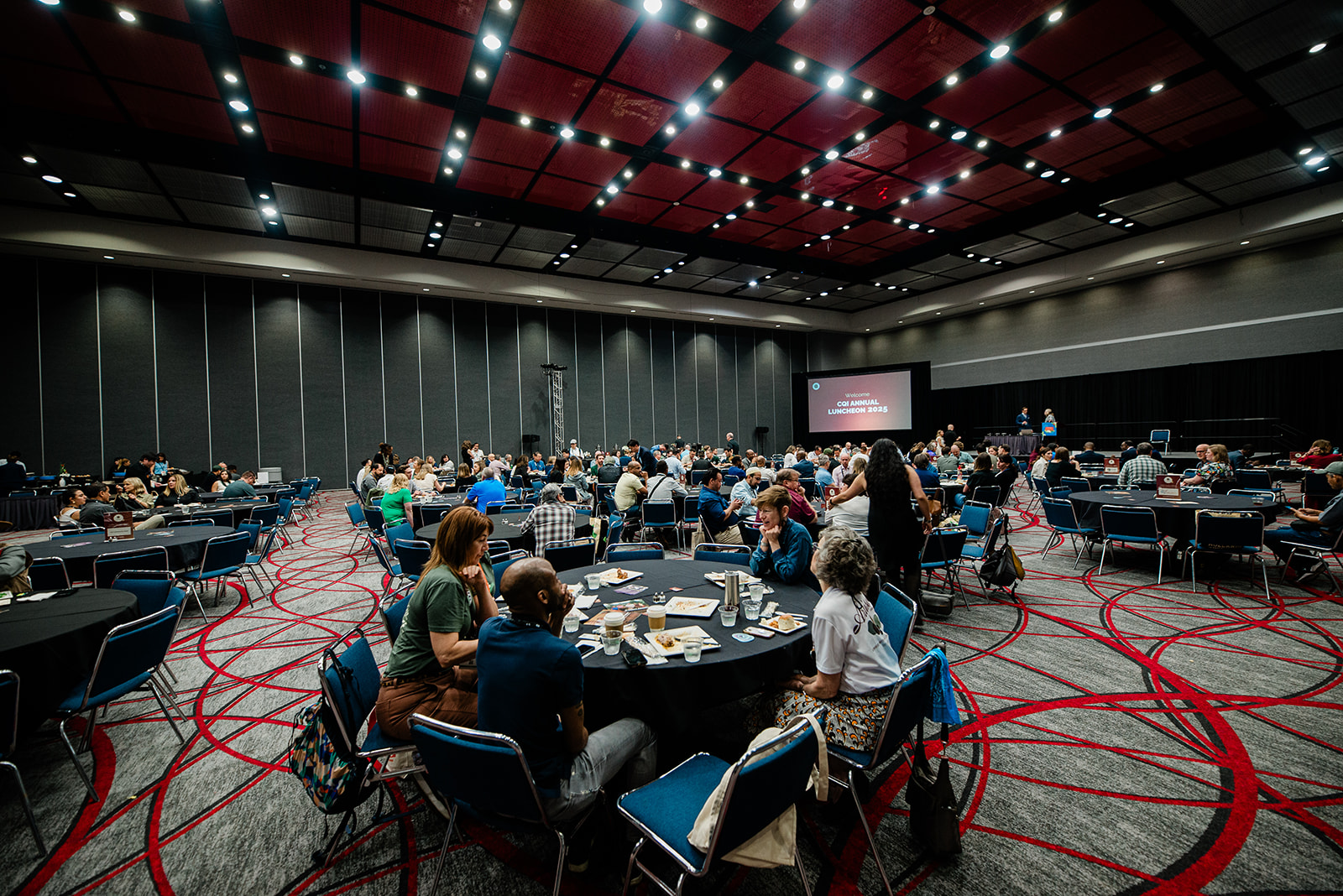 Conference room with people seated at round tables having a luncheon event, a screen in the background reads 'Welcome CQI Annual Luncheon 2025'.