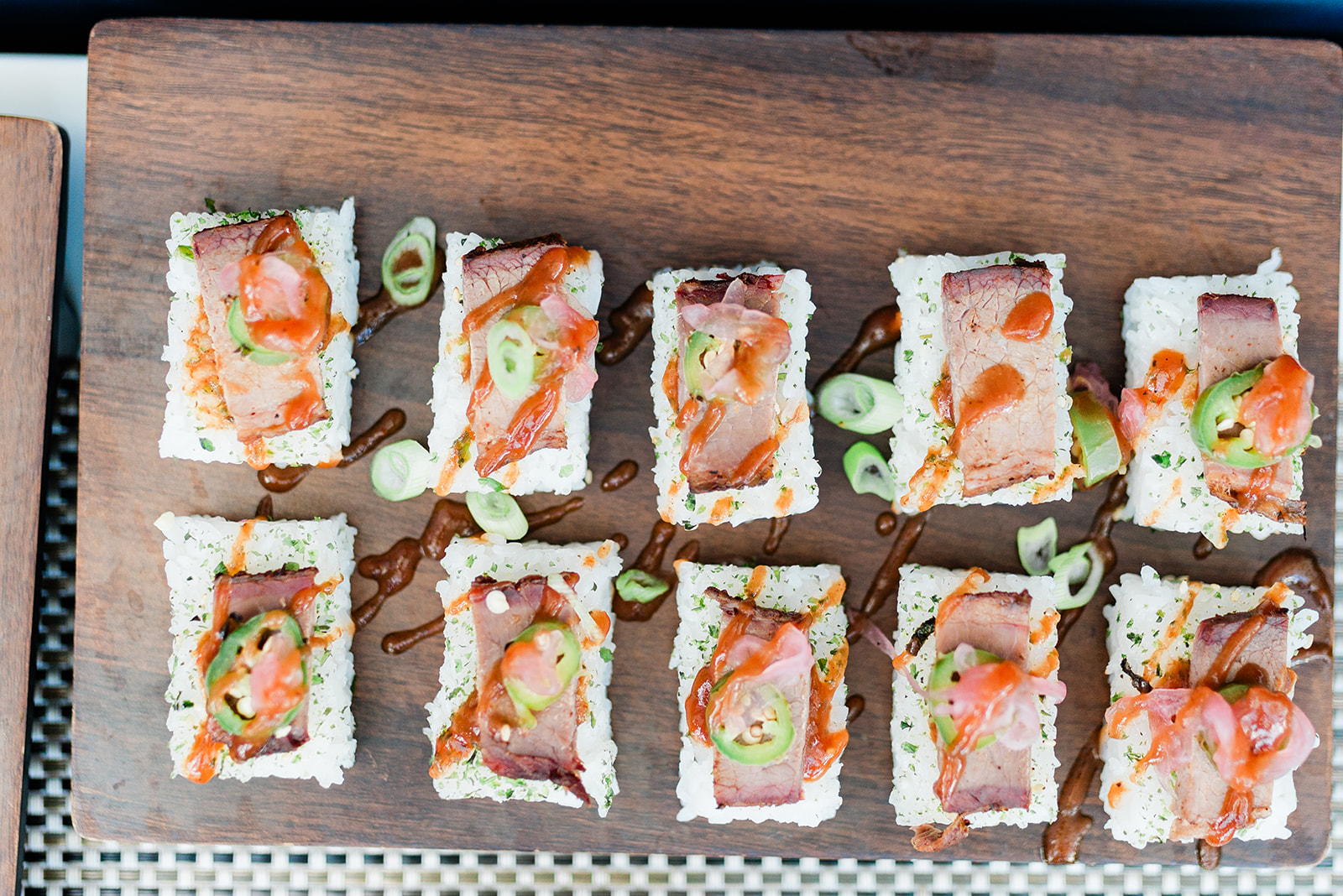Rectangular sushi pieces topped with seared meat, sliced jalapeños, pickled ginger, and sauce on a wooden serving board.