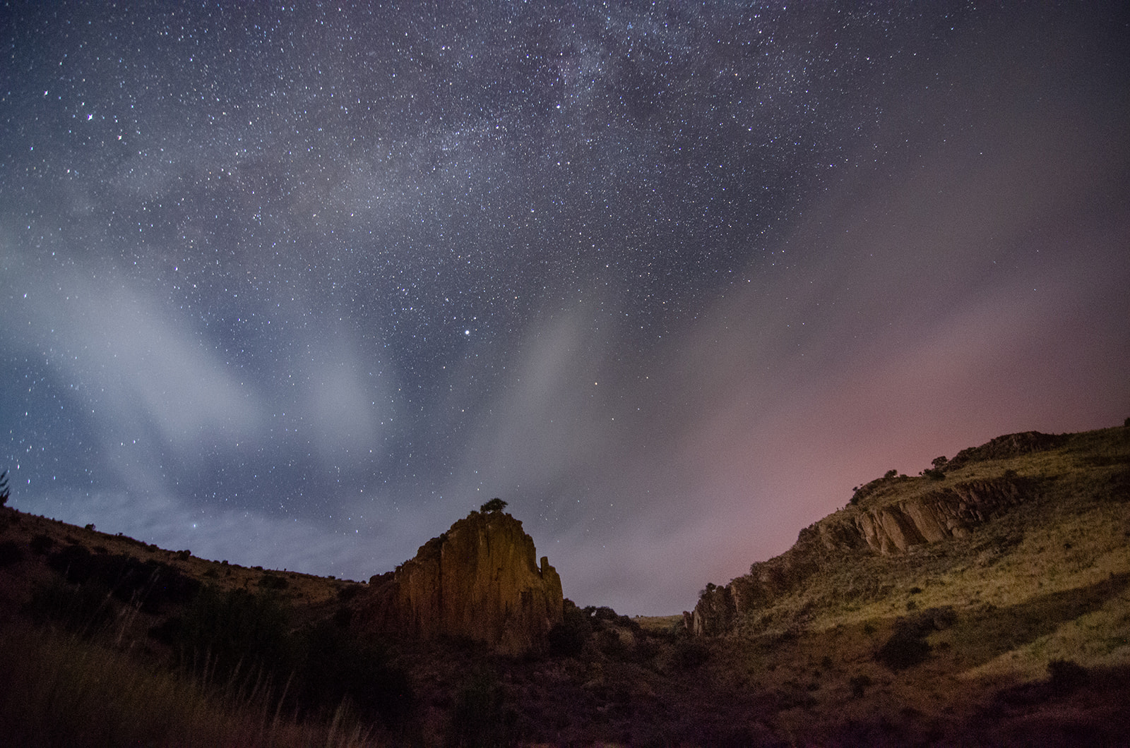 Starry night sky over rocky hills with light clouds and a faint pink glow on the horizon.