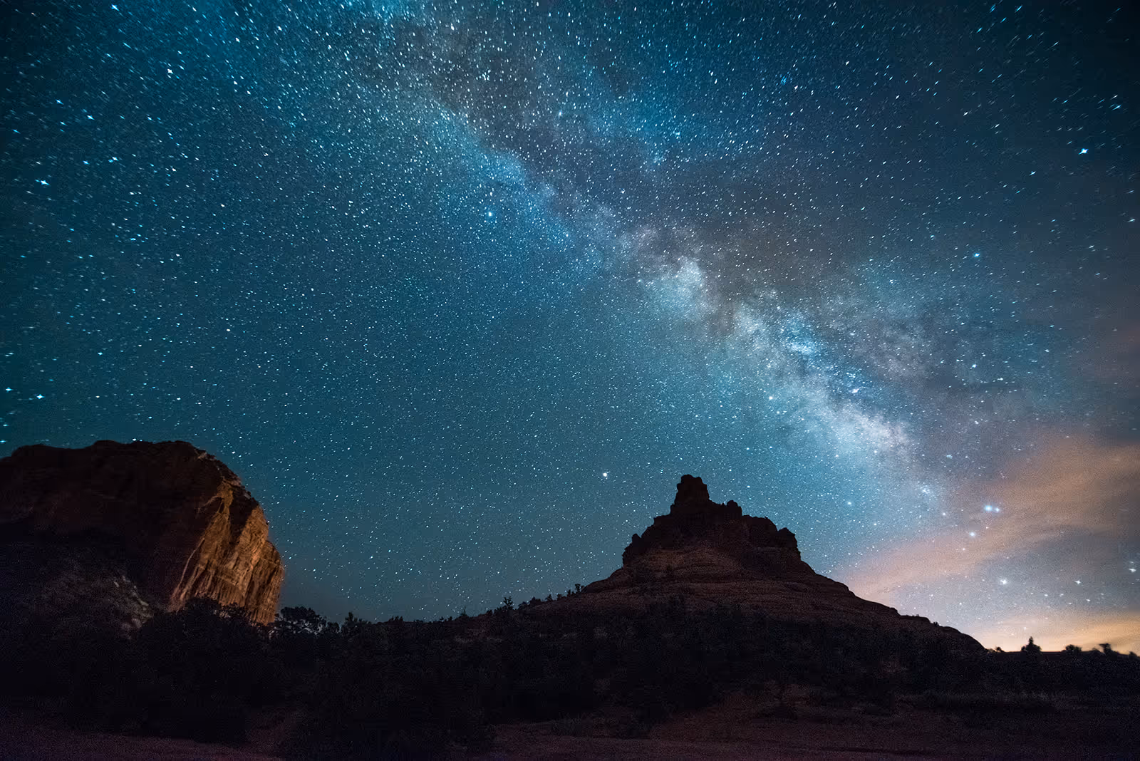 Night sky filled with stars and the Milky Way galaxy arching over dark silhouetted rock formations.