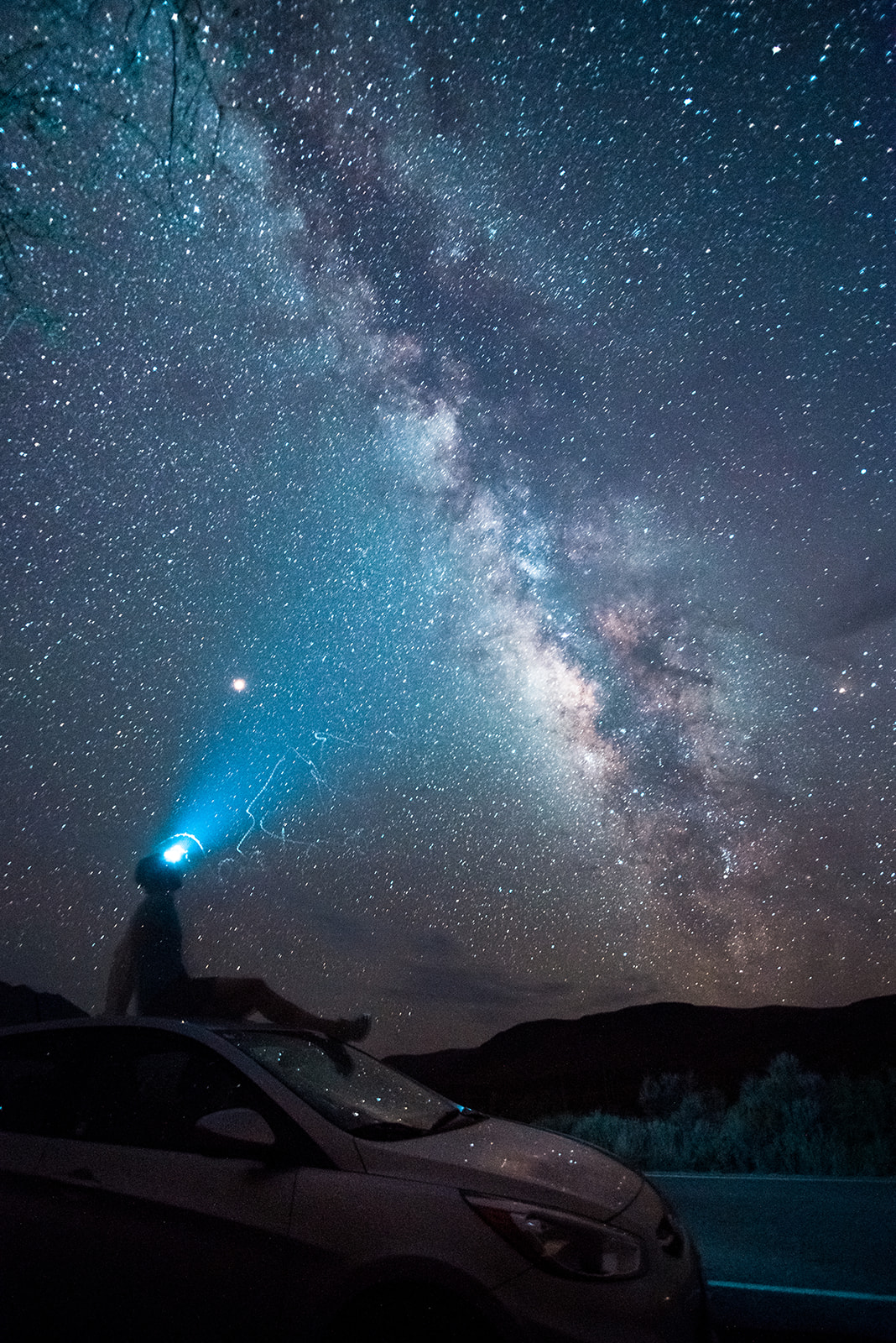 Person sitting on a car roof shining a blue headlamp into a starry night sky with the Milky Way visible.