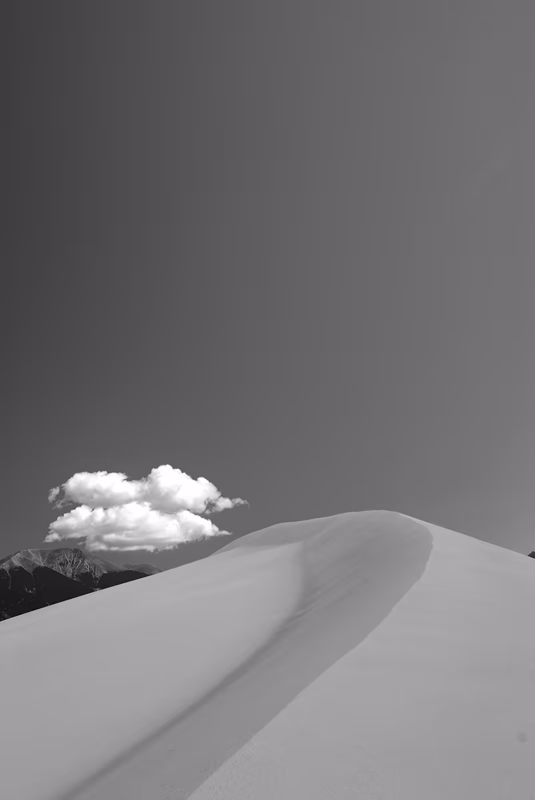 Smooth sand dune under clear sky with a single fluffy cloud and distant mountains in the background.