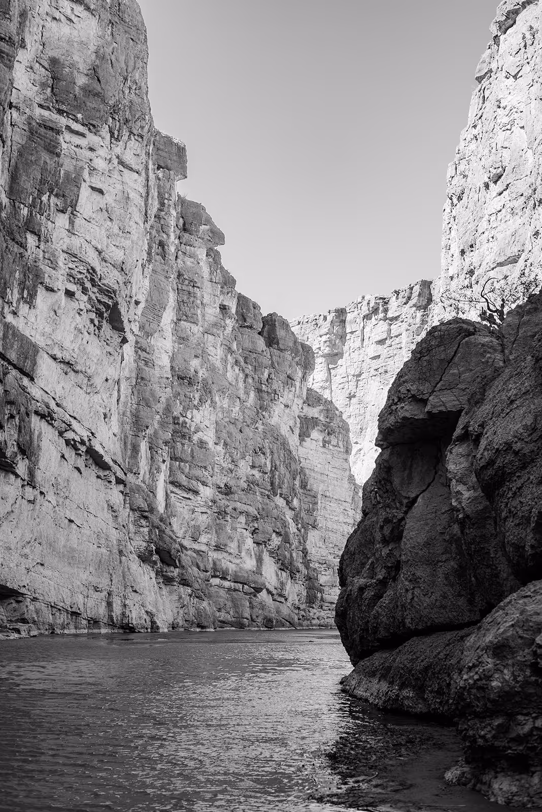 Black and white photo of a narrow canyon with steep rocky cliffs and calm water flowing through.