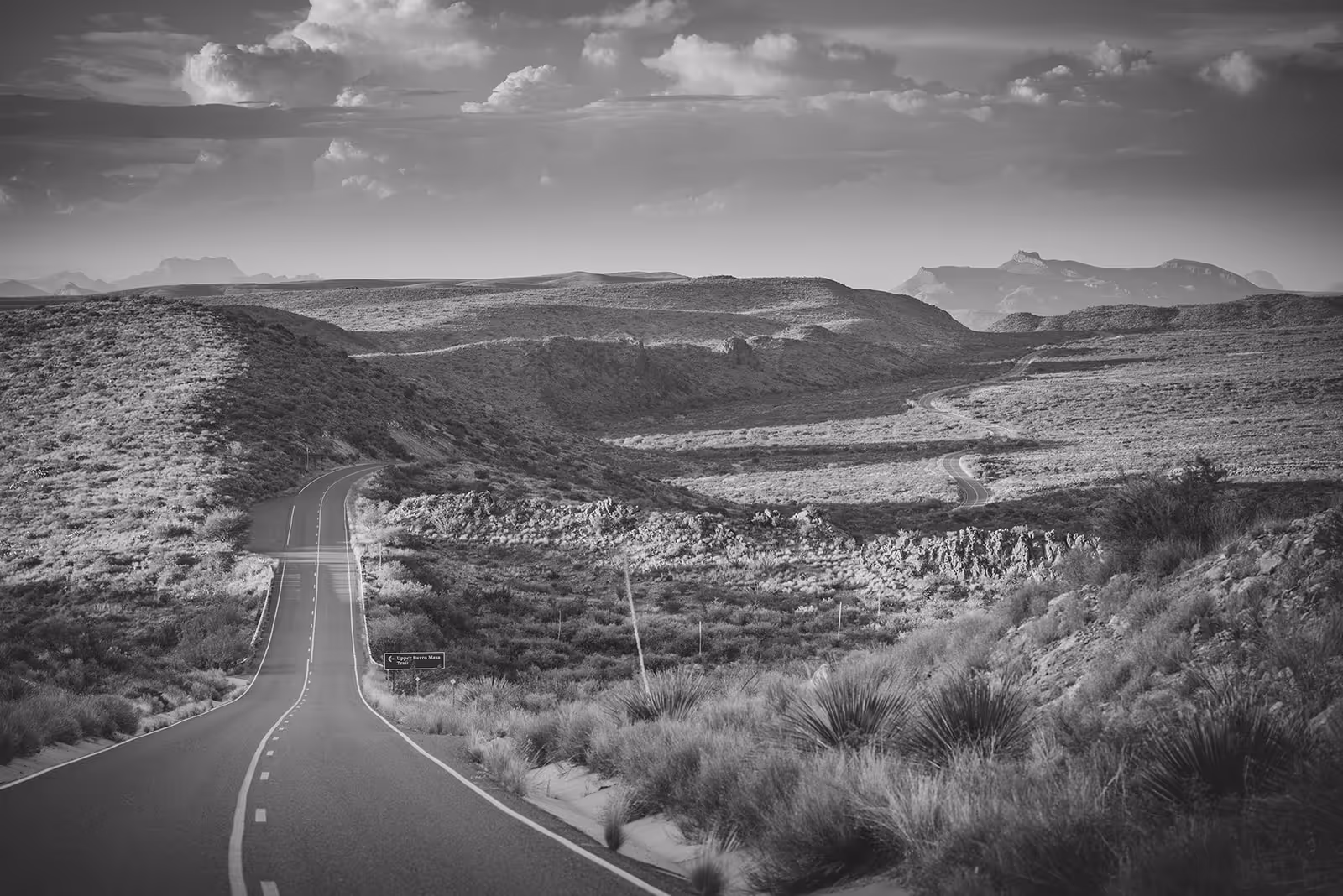 Winding road stretching through desert hills with distant mountains under a cloudy sky.