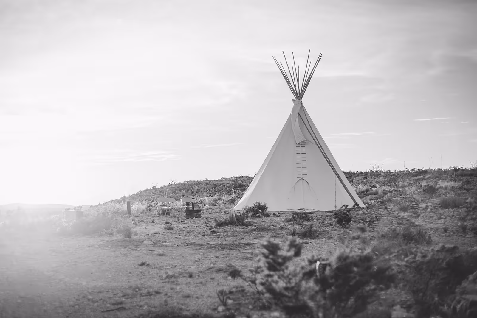 Black and white photo of a single teepee on a barren landscape with two folding chairs and a basket nearby.