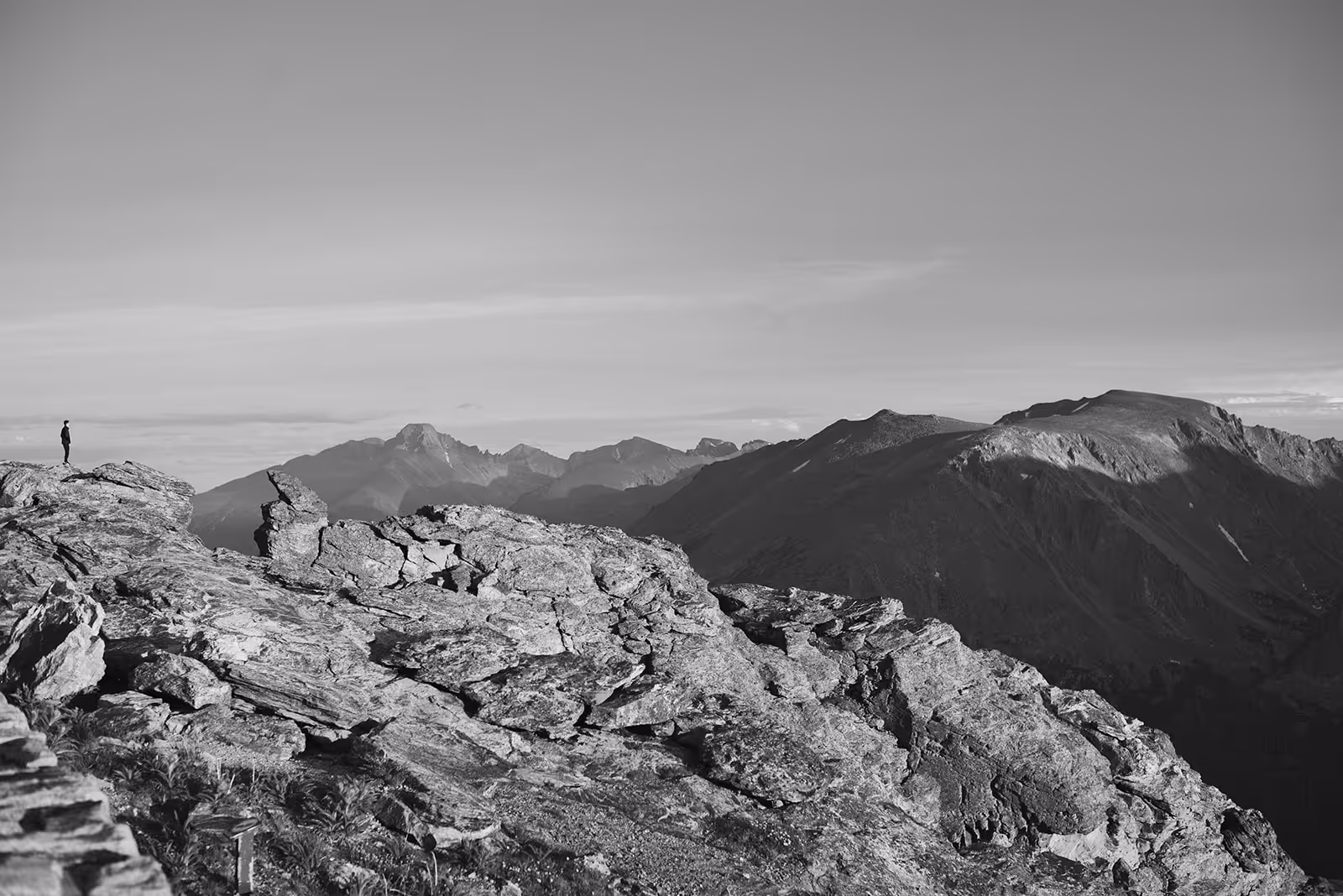 Person standing alone on rocky mountain peak overlooking a range of distant mountains under a clear sky.