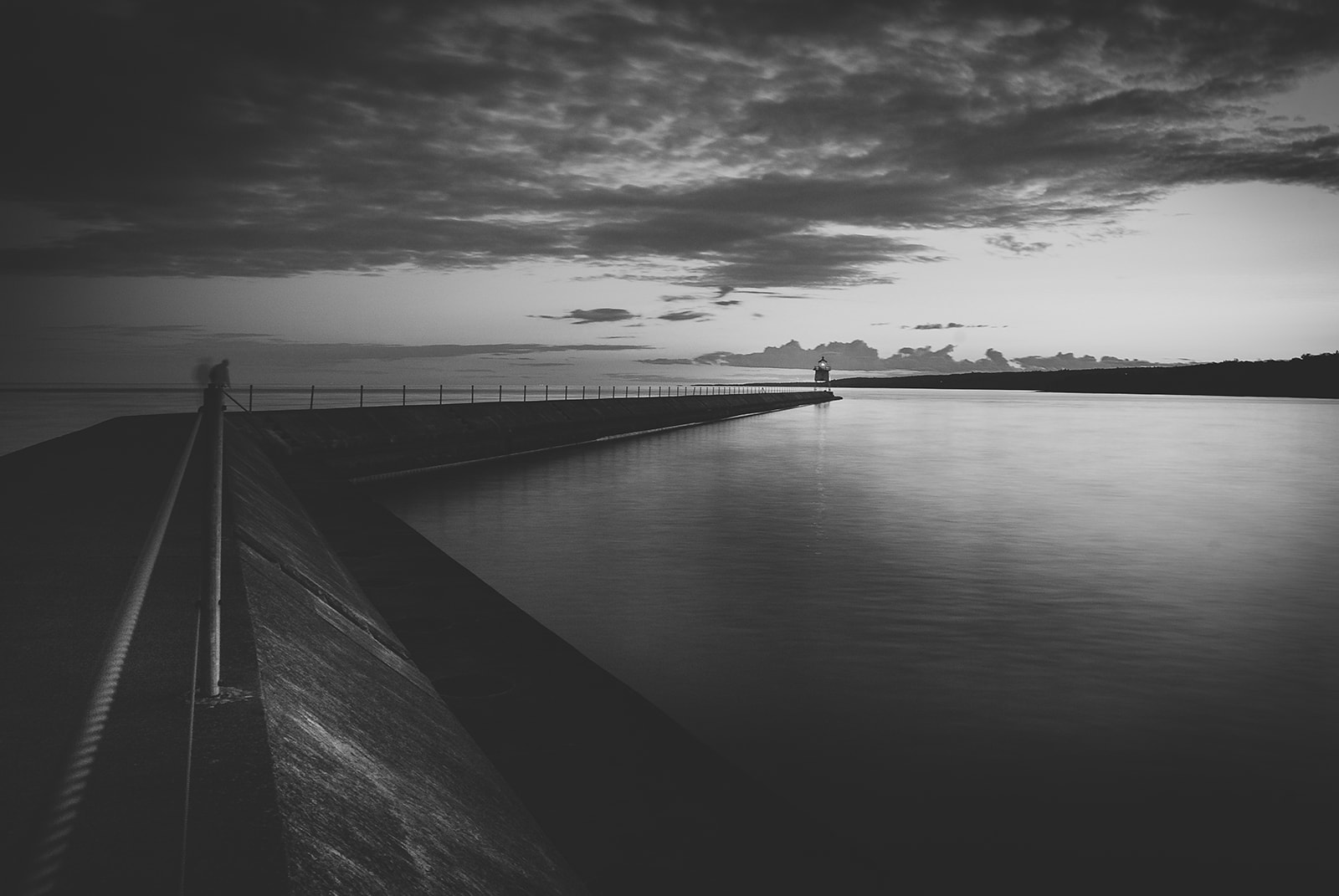 Long pier extending into calm water with a lighthouse at the end under a cloudy sky at dusk.