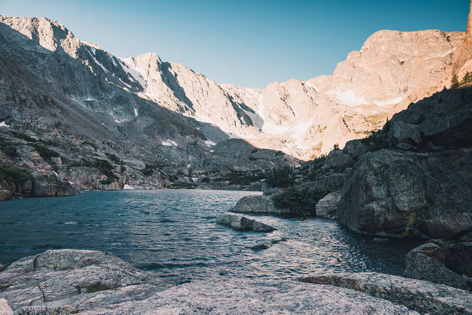 Mountain lake surrounded by rocky cliffs with patches of snow under a clear blue sky.