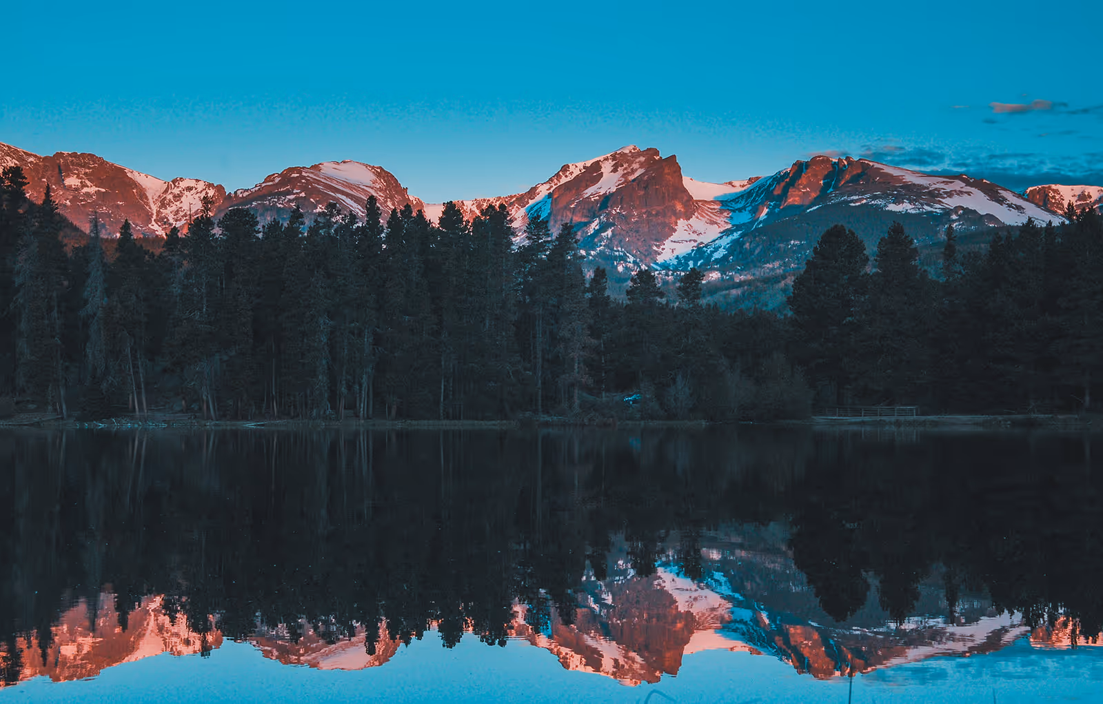 Snow-capped mountain range glowing pink at sunset reflected on a still lake with a dense dark pine forest along the shore.