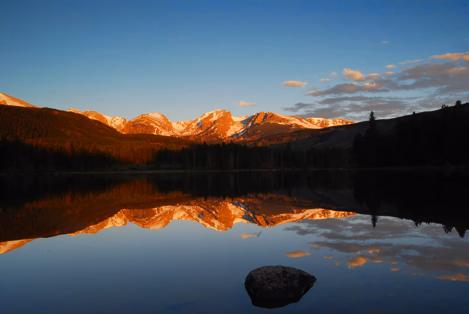 Snow-capped mountains at sunset reflecting on a calm lake with a rock in the foreground.