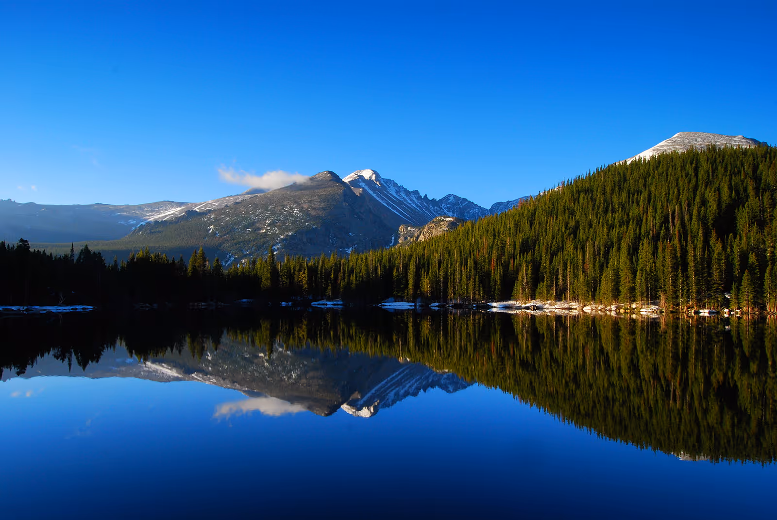 Clear mountain lake reflecting forested slopes and snow-capped peaks under a bright blue sky.