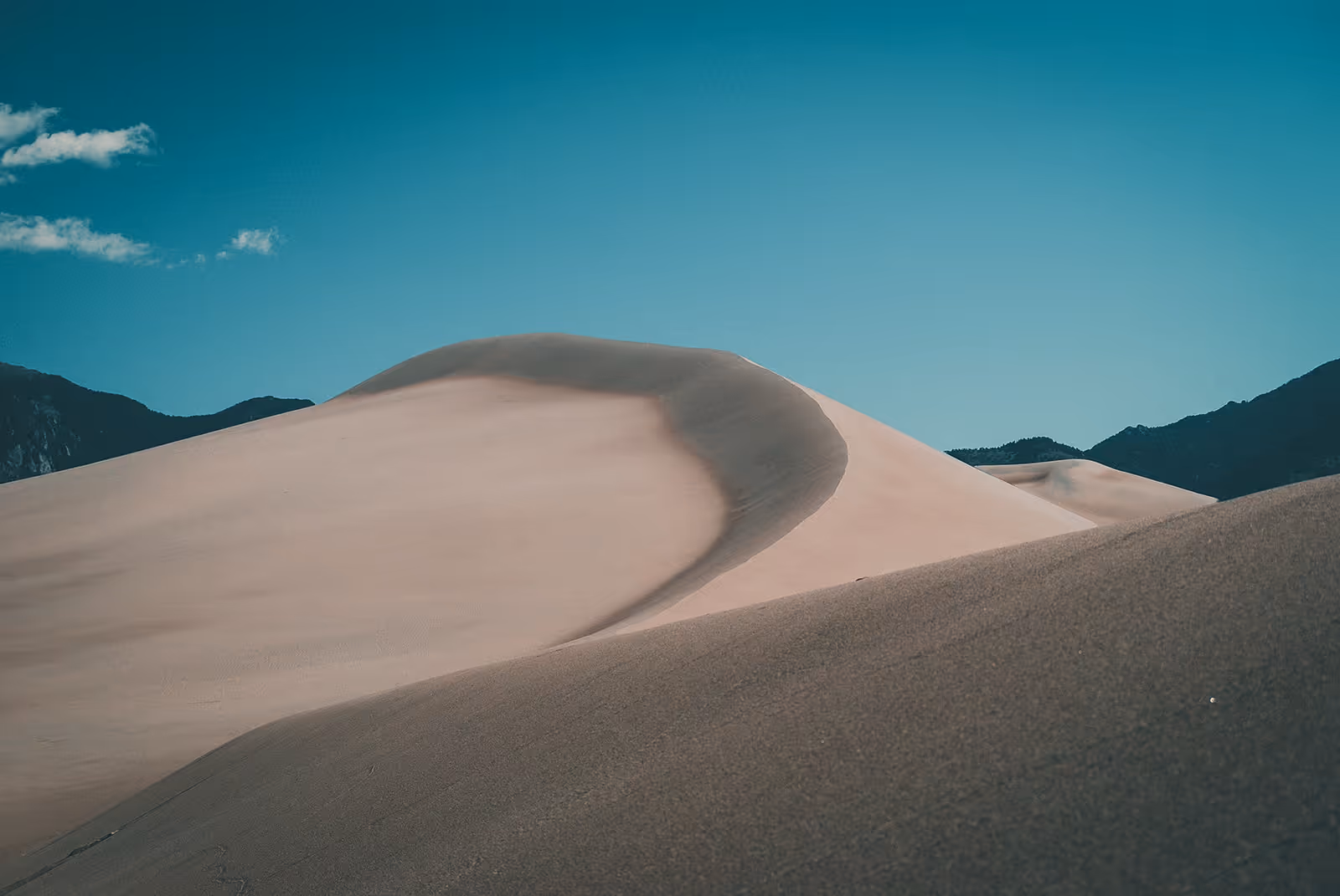 Large sand dunes under a clear blue sky with mountains in the background.