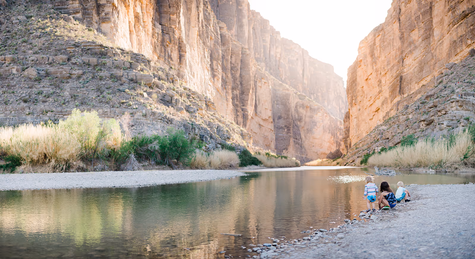 Three children playing by the edge of a calm river with towering canyon cliffs and shrubs in the background.
