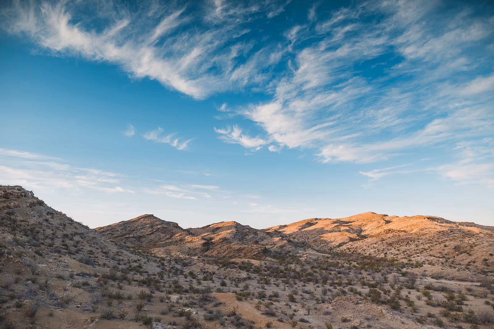 Desert mountains under a blue sky with wispy clouds during golden hour.