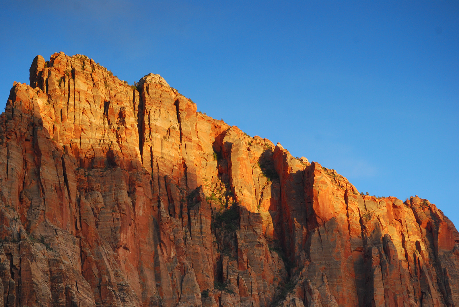 Sunlit red rock cliff with vertical striations under a clear blue sky.