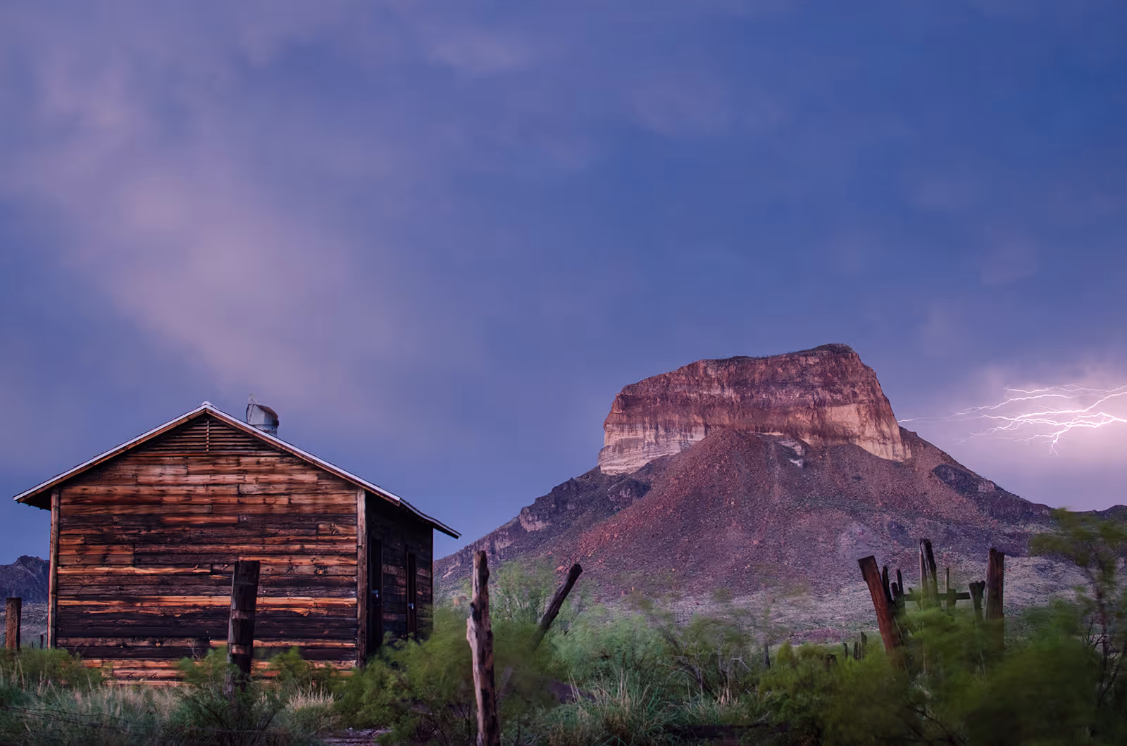 Wooden cabin in a grassy area before a rocky mountain with lightning in a purple sky.
