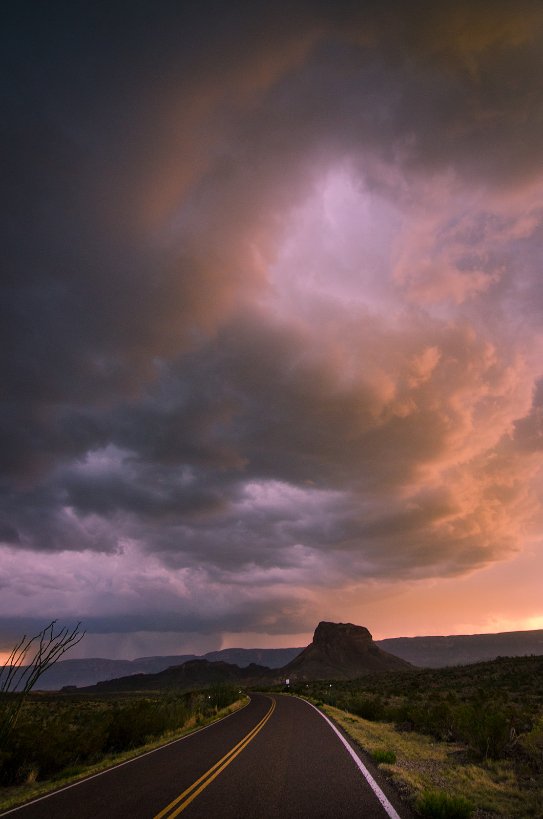 Empty road leading toward a mesa under dramatic purple and orange storm clouds at sunset.
