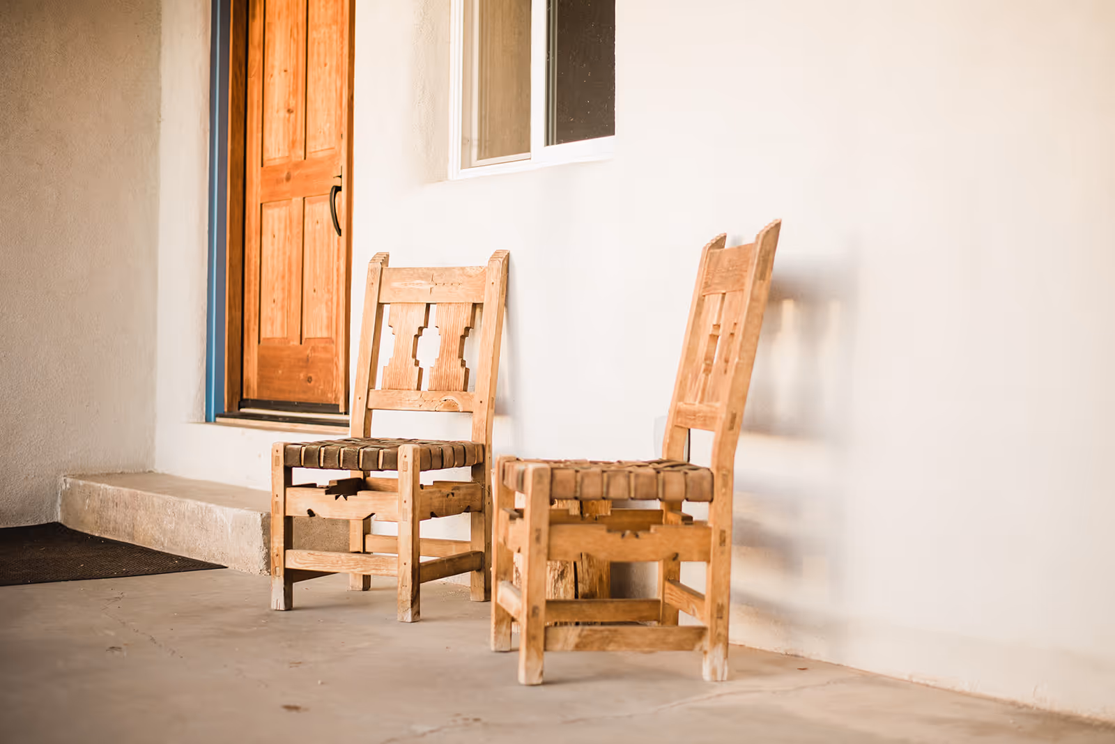 Two rustic wooden chairs with woven seats on a beige concrete porch next to a wooden door and window.