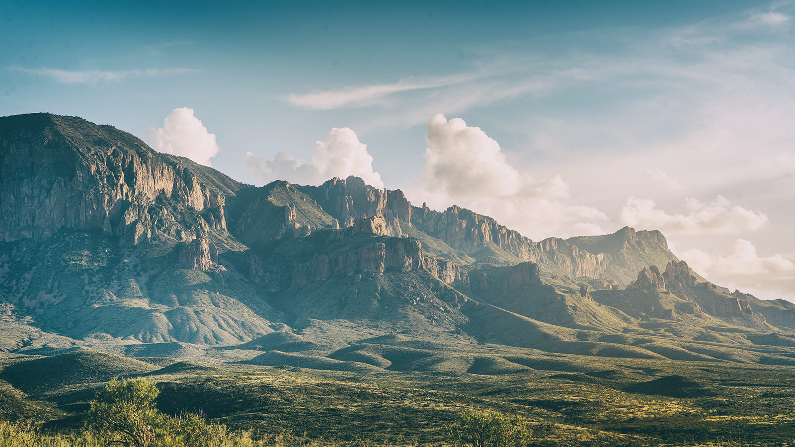 Sunlit rugged mountain range with rocky cliffs under a partly cloudy blue sky.