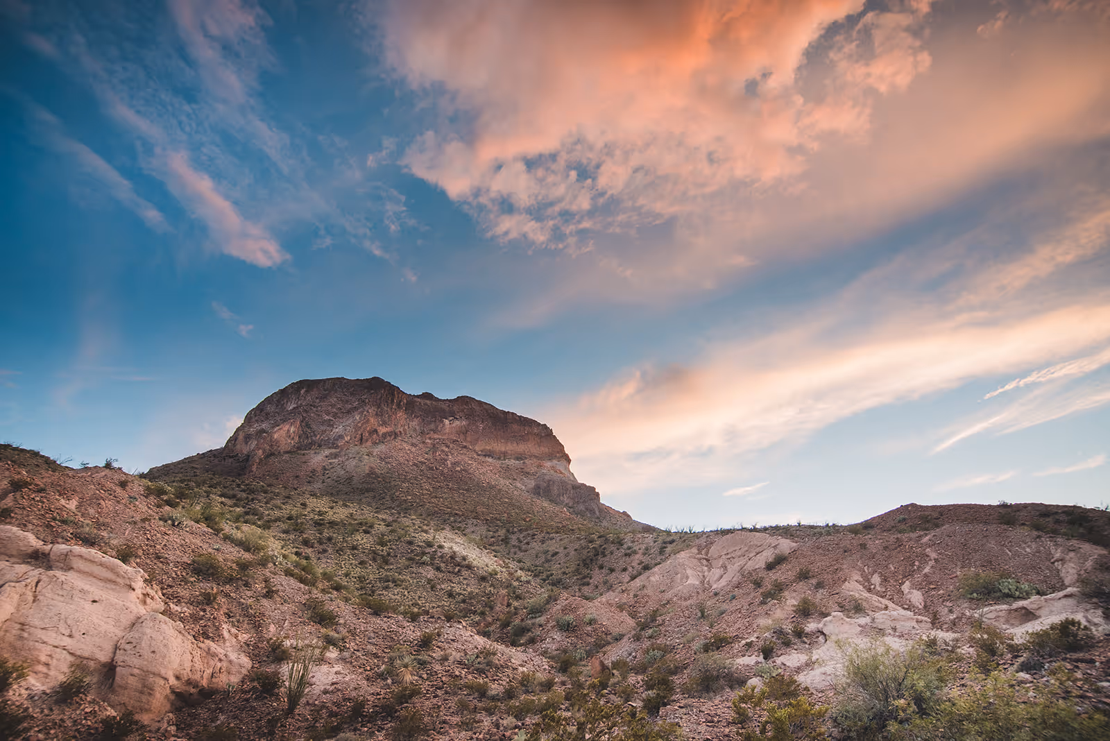 Rocky desert mountain under a blue sky with pink and orange clouds at sunset.