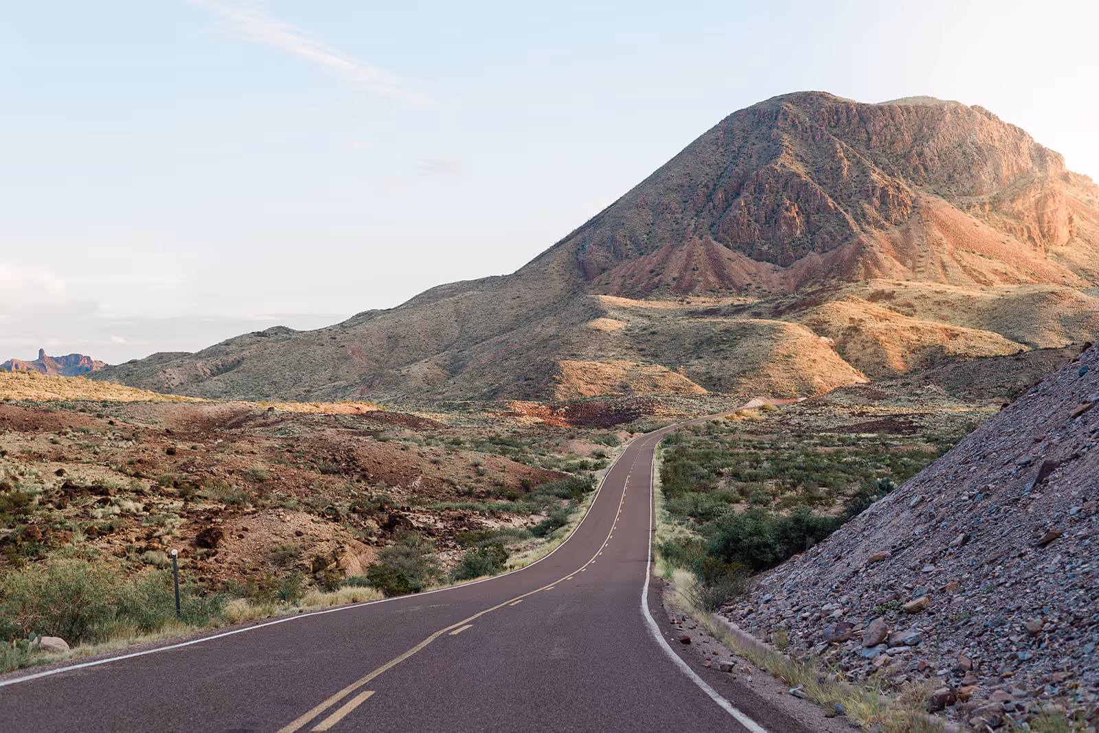 Two-lane road winding through rocky desert landscape with a large mountain in the background under a clear sky.