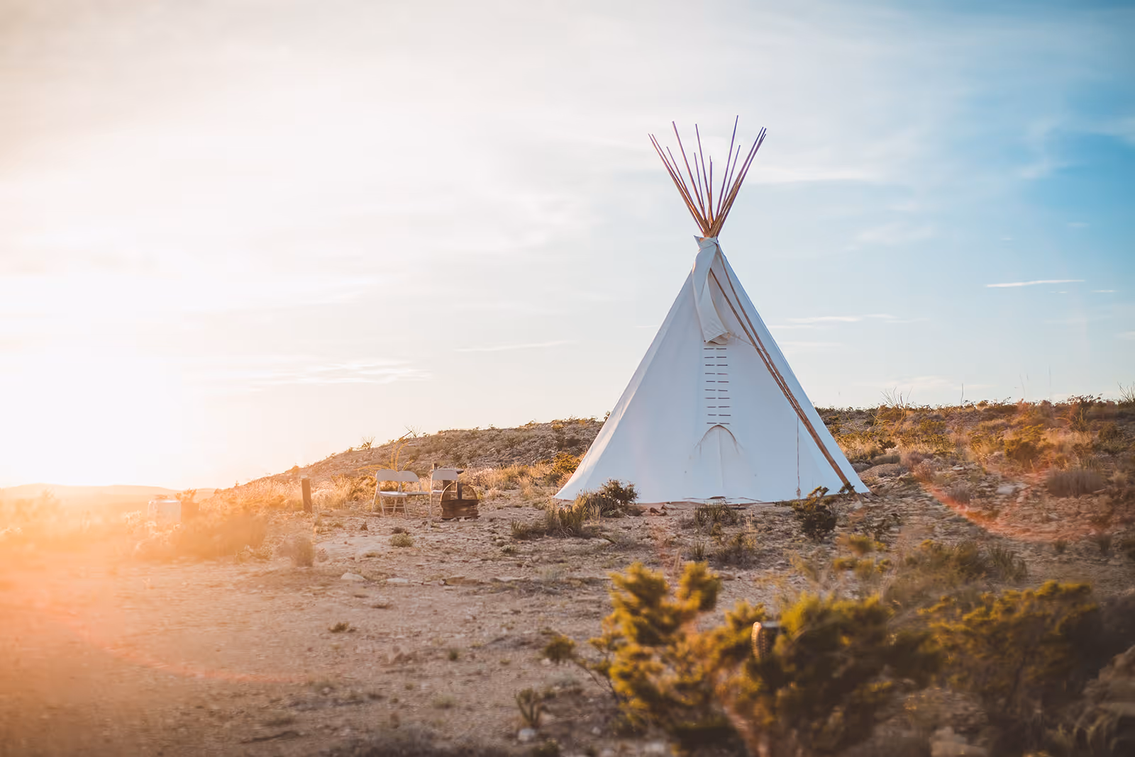 White teepee tent set up on rocky terrain with two folding chairs and a fire pit at sunset.