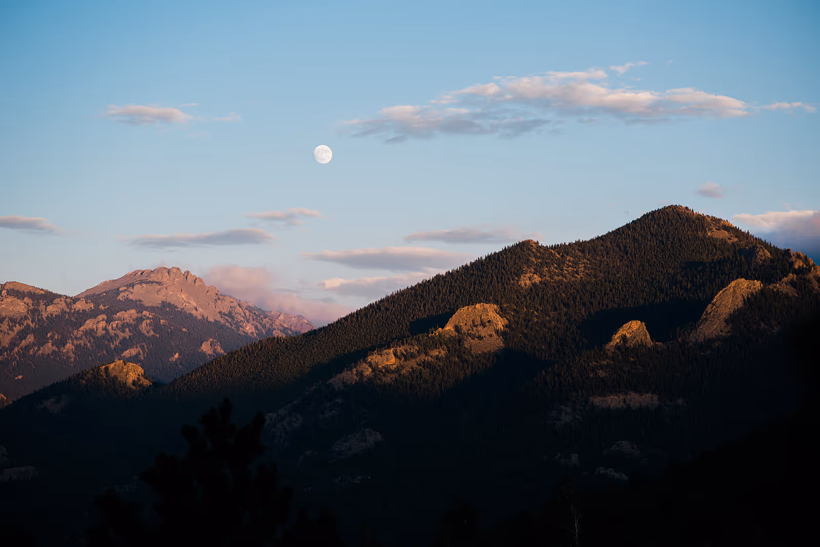 Mountain range with tree-covered peaks illuminated by sunset light under a clear sky with a visible full moon.