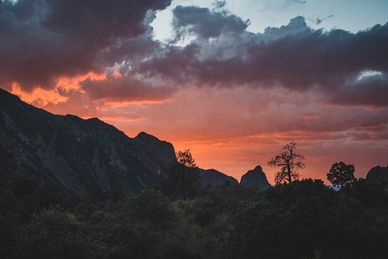 Sunset casting an orange glow behind silhouetted mountains and trees with dark clouds overhead.