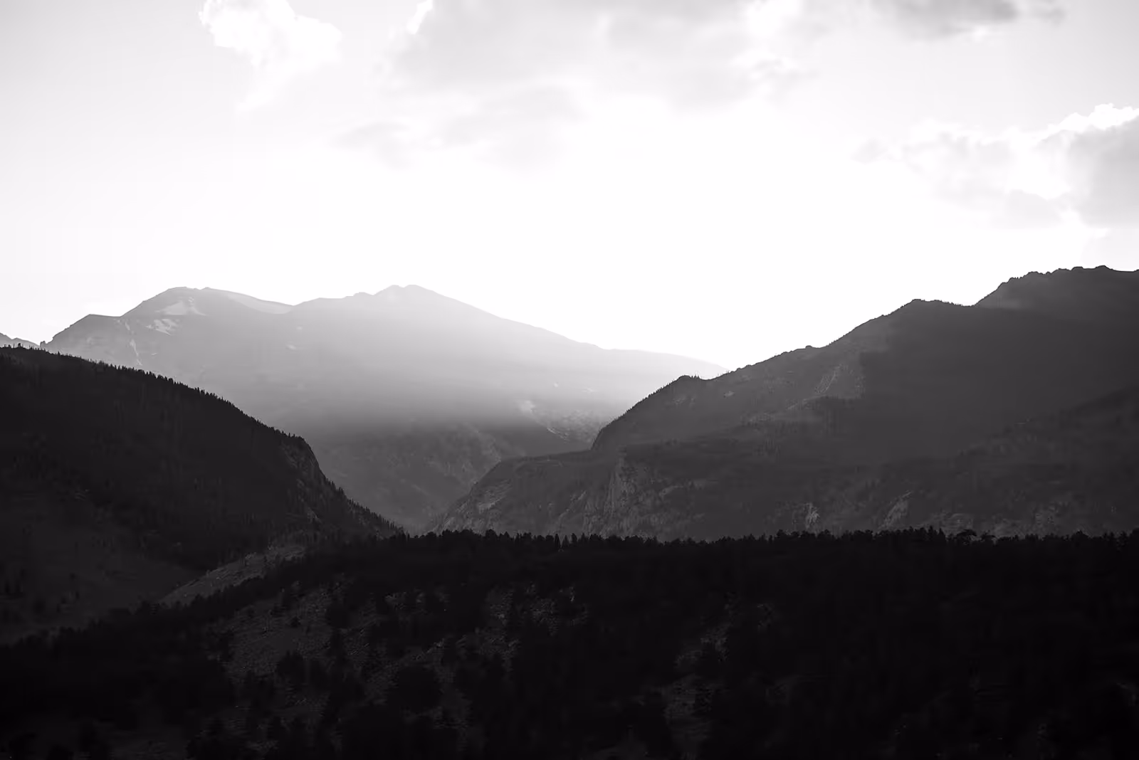 Black and white photo of mountain ranges with sunlight breaking through clouds behind the peaks.
