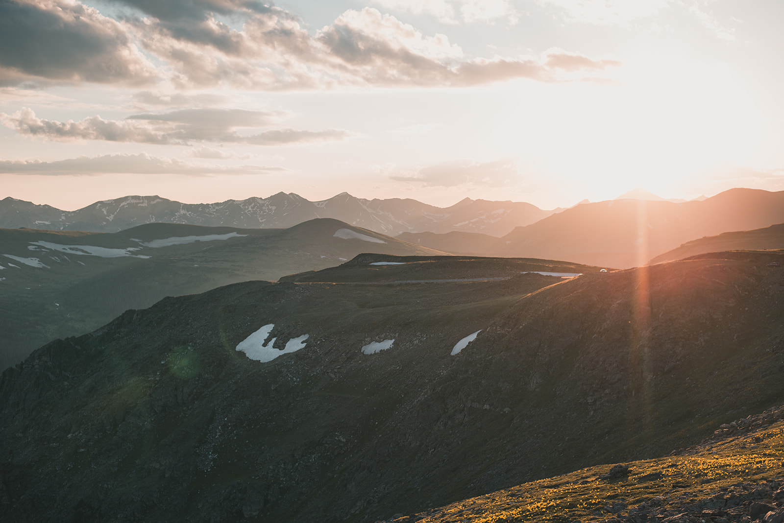 Sunset over a mountain ridge with patches of snow and distant snow-capped peaks under a partly cloudy sky.