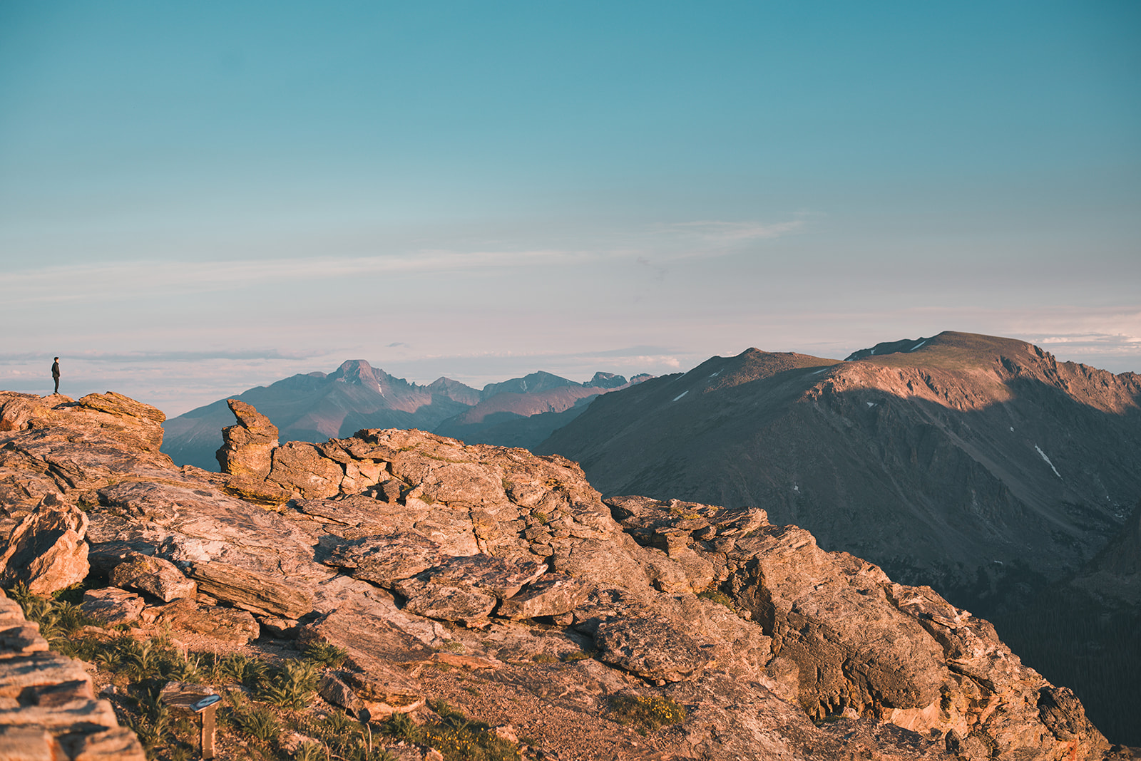 Person standing on rocky mountain ledge overlooking distant sunlit mountain range under clear blue sky.