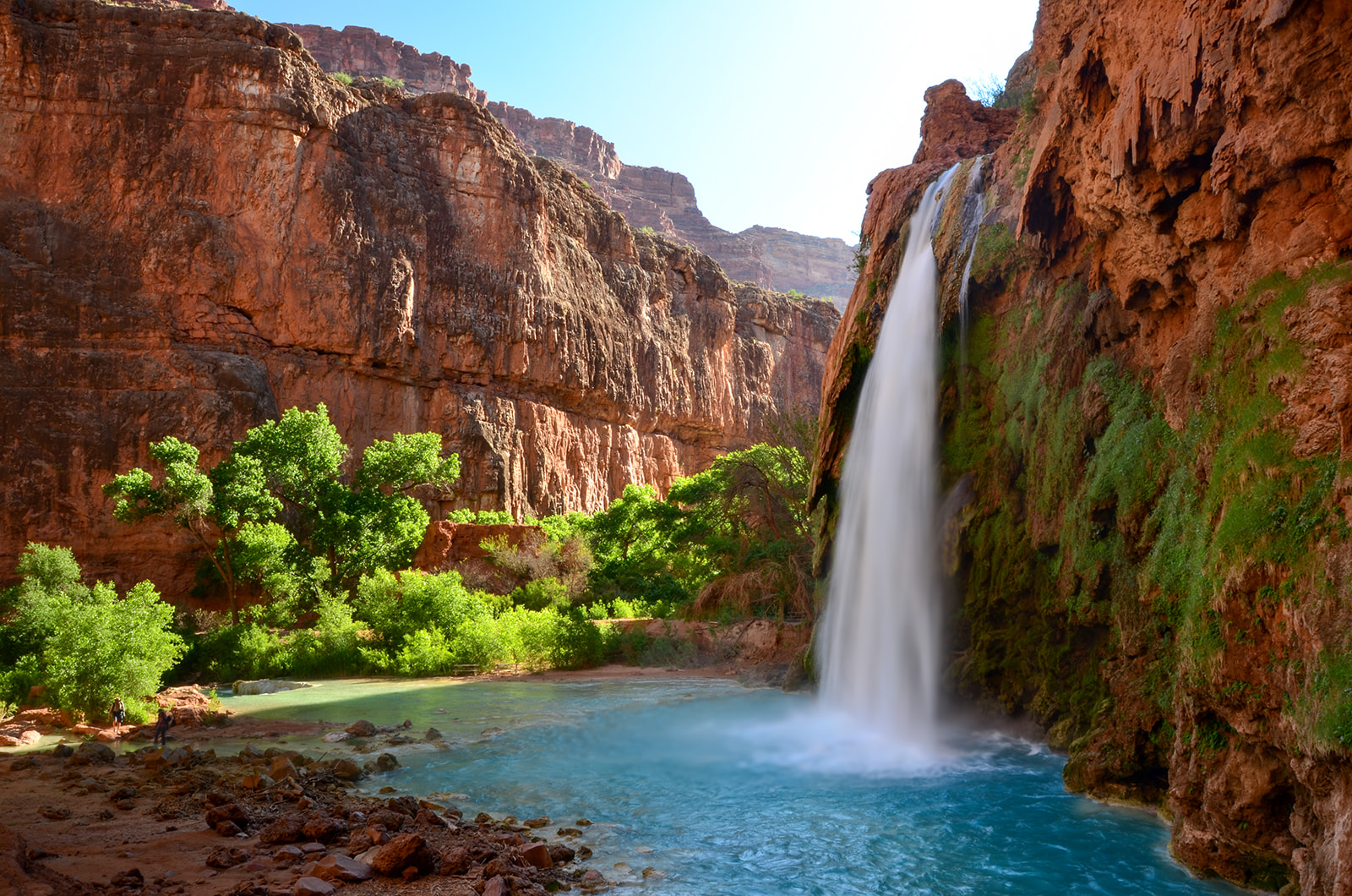 Waterfall cascading into a blue pool surrounded by red rock cliffs and green trees under a clear sky.