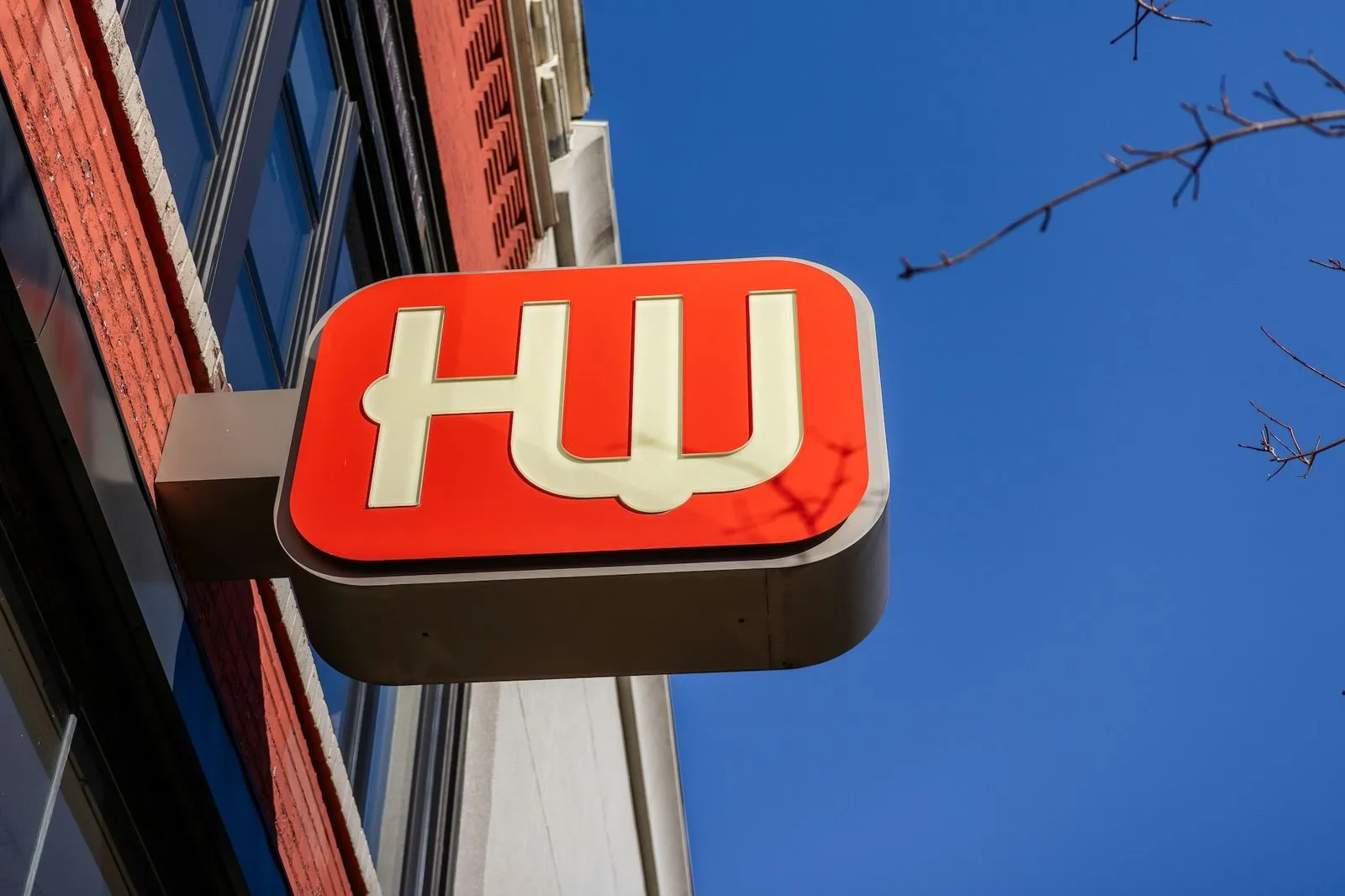 HW logo sign mounted on a building wall under a clear blue sky with tree branches nearby.