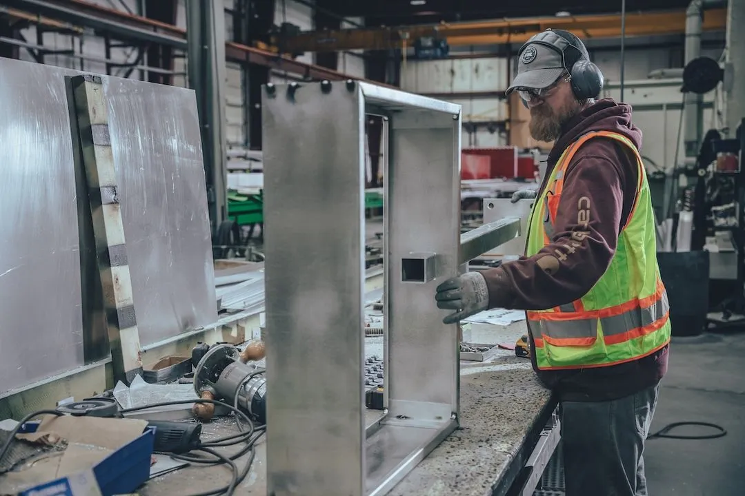 Worker in safety vest and ear protection handling a large metal frame in an industrial workshop.