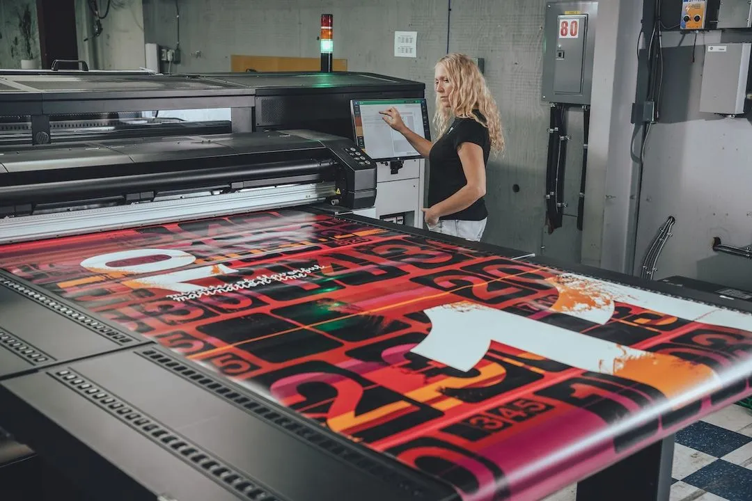 Woman operating a large-format printer producing a colorful graphic print with red, black, and orange tones.