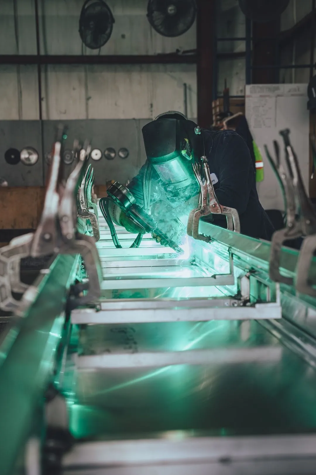 Welder wearing protective gear fusing metal pieces in an industrial workshop with green welding light.