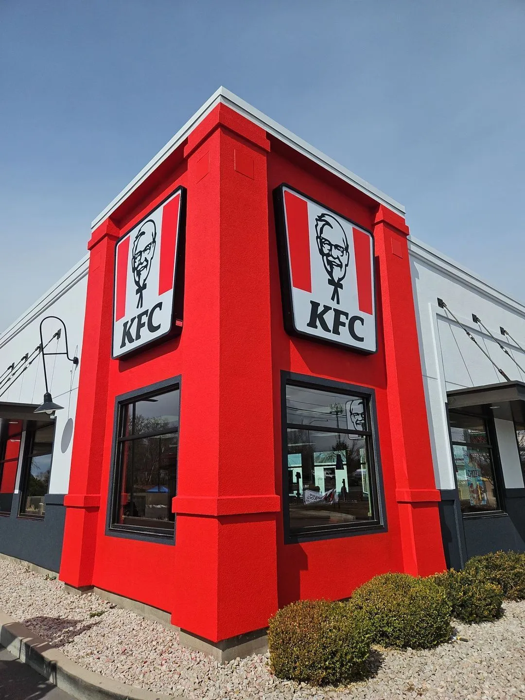 Corner view of a KFC restaurant with bright red exterior walls, large windows, and KFC logo signs featuring Colonel Sanders.