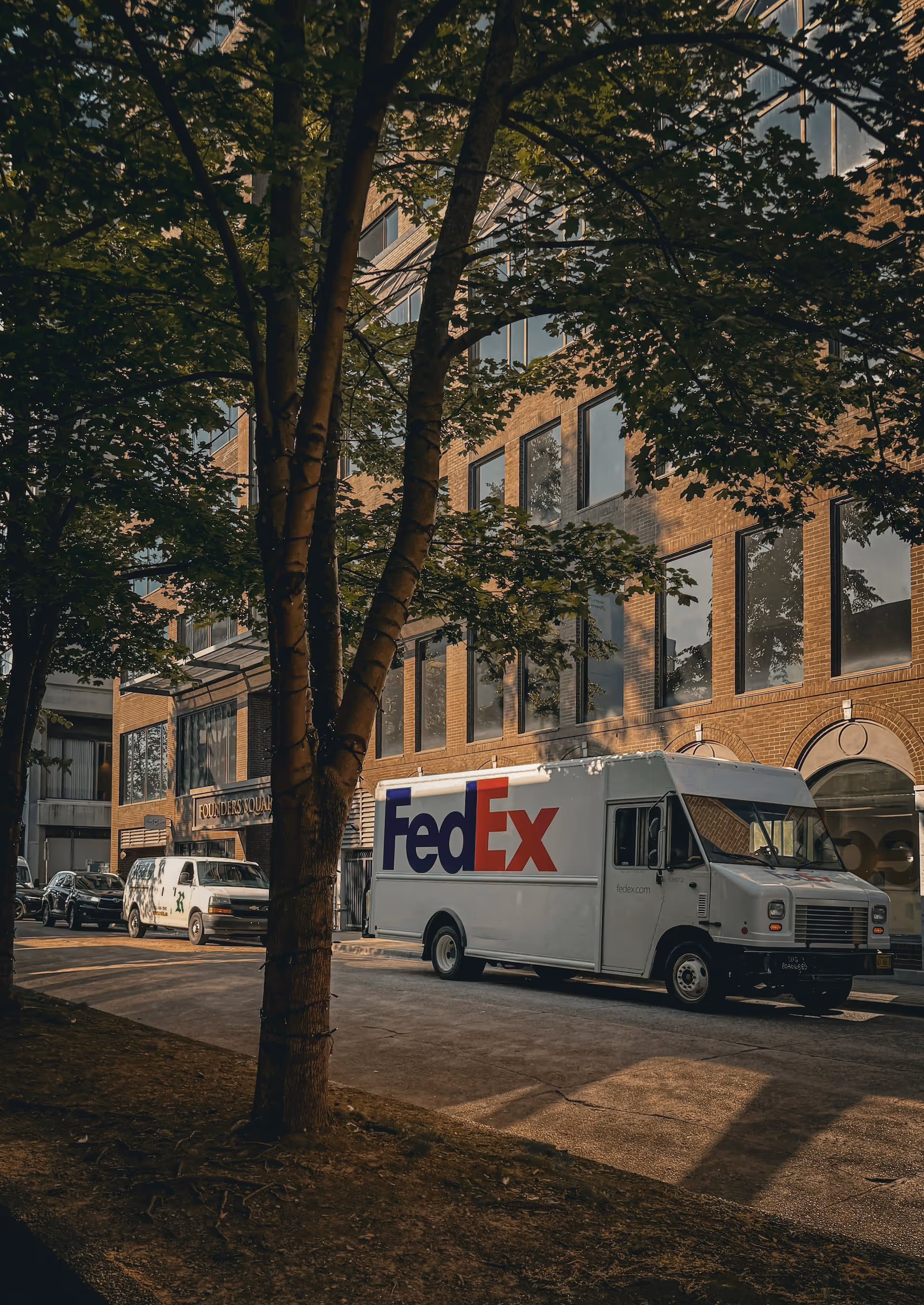 FedEx delivery truck parked on a city street next to a brick building with large windows, partially obscured by tree branches.
