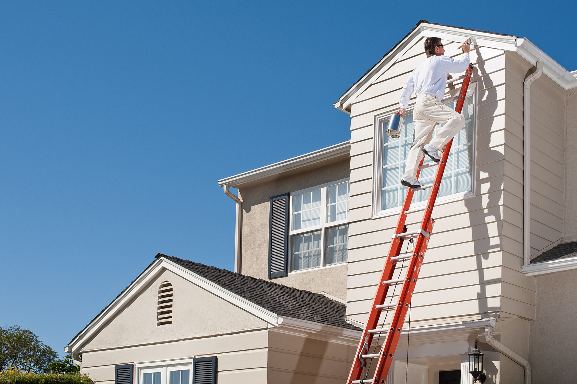 Worker painting trim white beneath a roof of a house with an orange extension ladder.