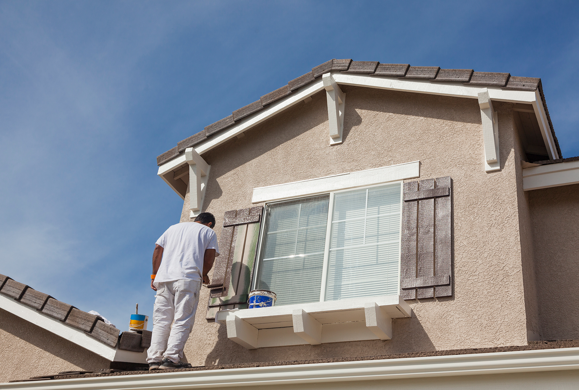 Worker painting shutters brown on the outside of a house.
