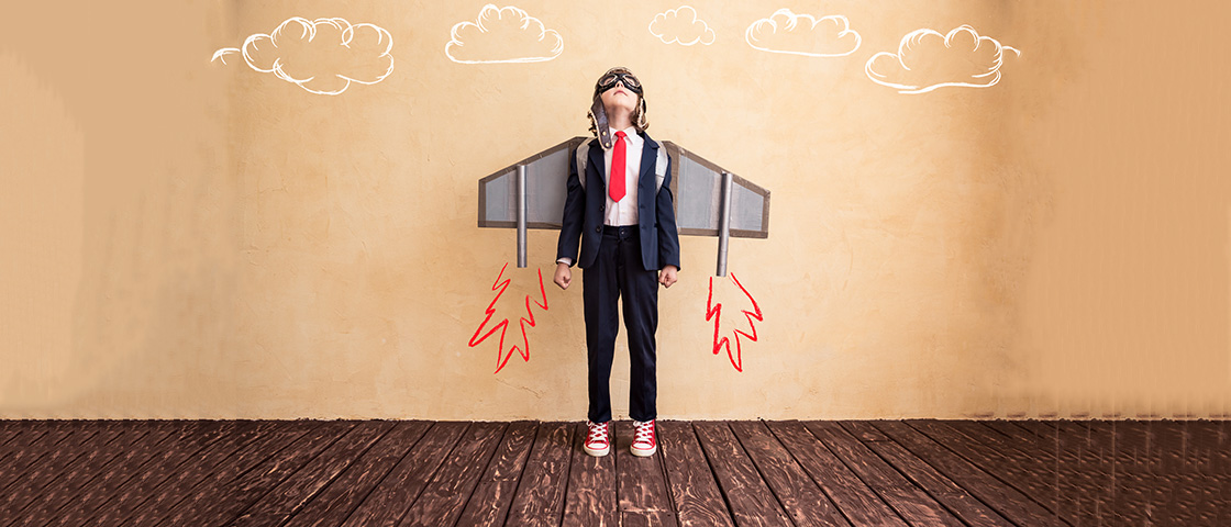 A young boy wearing pilot goggles and standing in front of a wall painted with rocket boosters, symbolizing student empowerment and imagination.