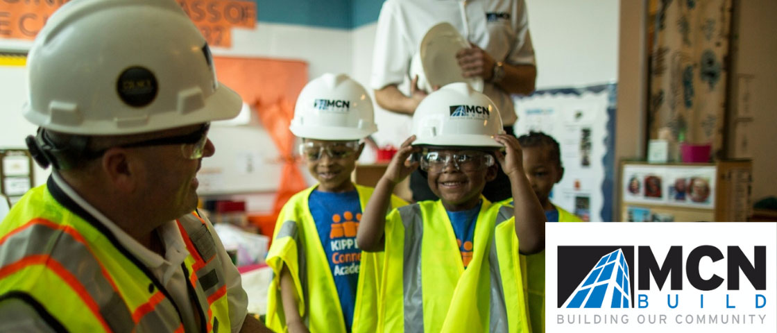 Children wearing safety glasses and yellow vests, standing with an adult in a safety vest and glasses, learning on a construction site, with the MCN Build logo overlayed.