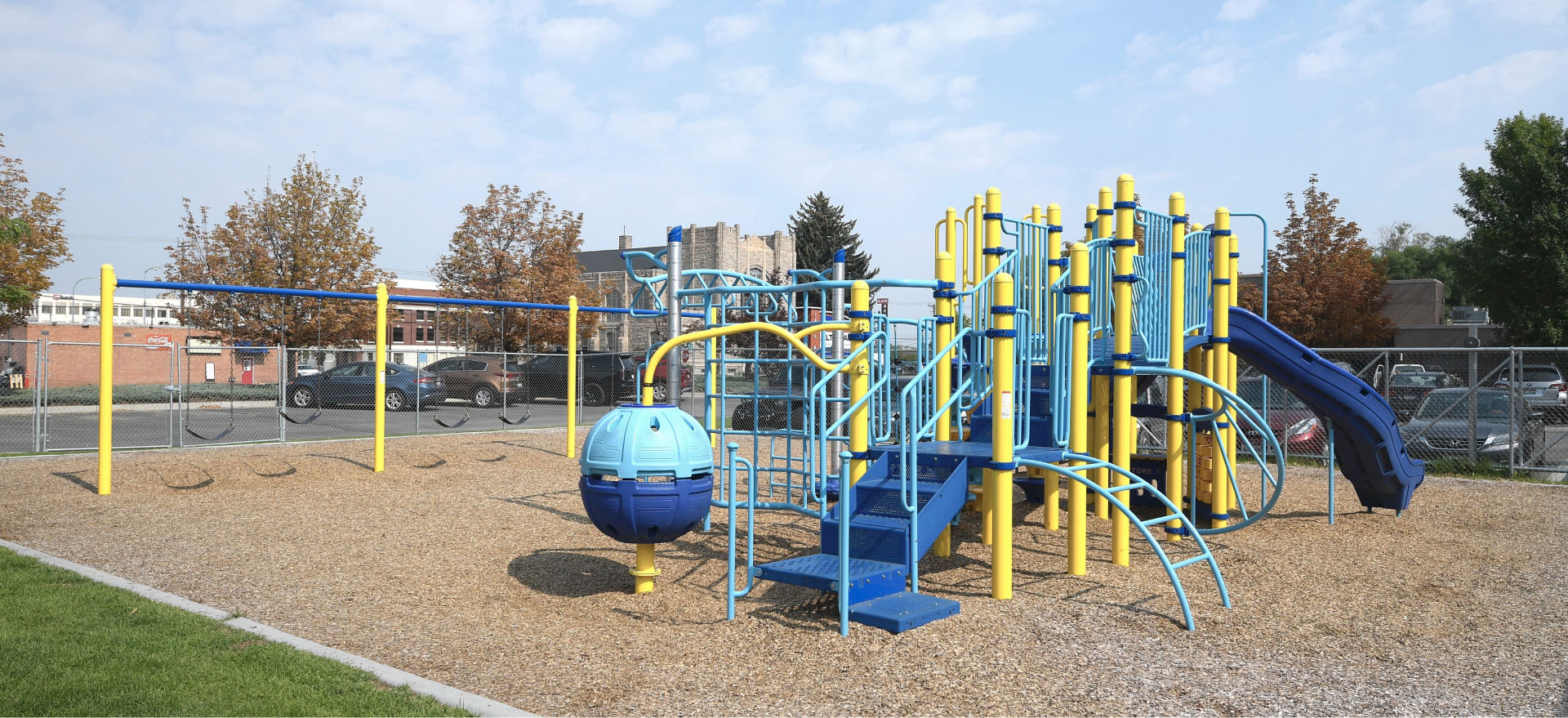 Accessible playground with blue and yellow climbing structures, slide, and swings in an open schoolyard.