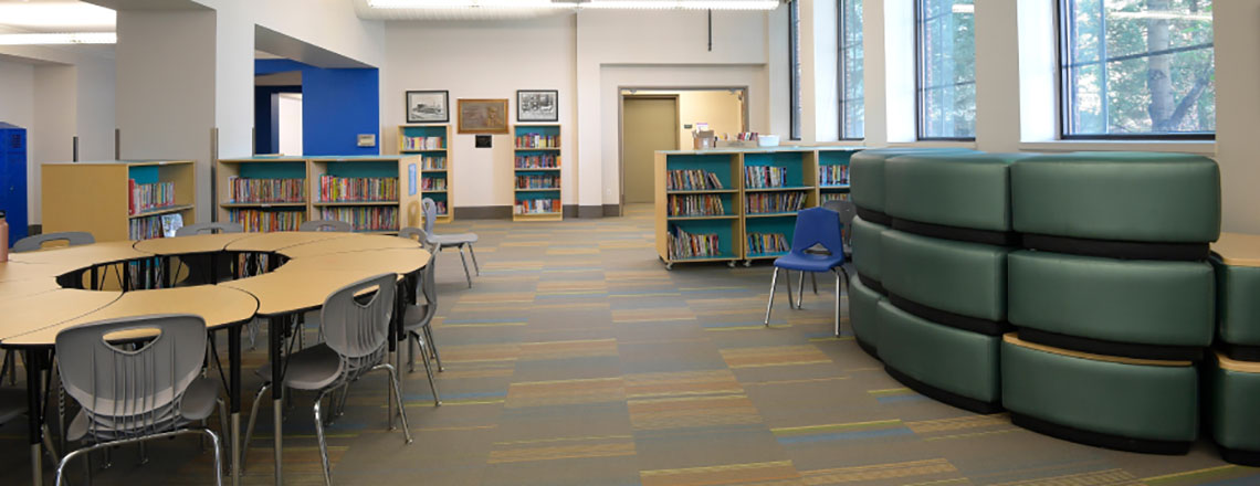 Modern school library with bookshelves, group tables, chairs, and flexible seating near large windows.