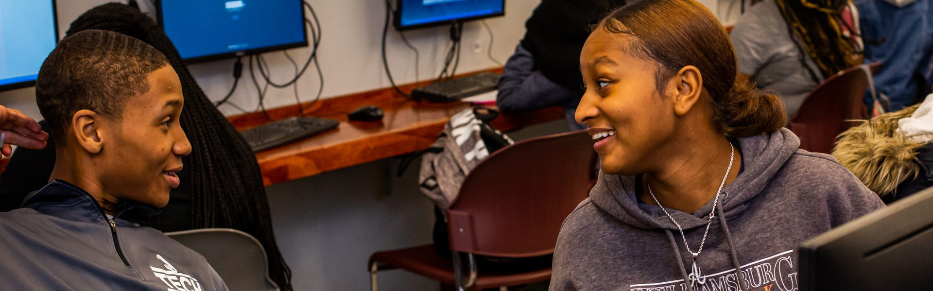 Two students smiling and talking in a computer lab with desktop screens behind them.